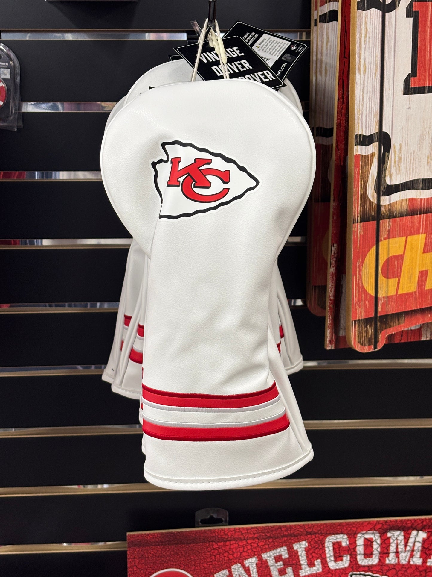 White golf club head covers with red accents and a logo on a shelf with sports team merchandise.